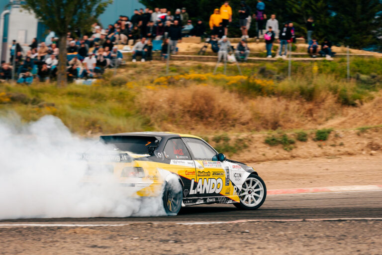 BMW E36 di Landoracing in pieno drift durante una gara, con fumo di gomme e pubblico sullo sfondo - Spettacolo di drifting adrenalinico.